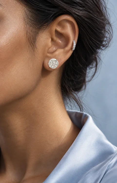 Close-up of a woman wearing a multi-stone diamond floral cluster stud and a small white-gold diamond huggie, with a pale blue collar.
