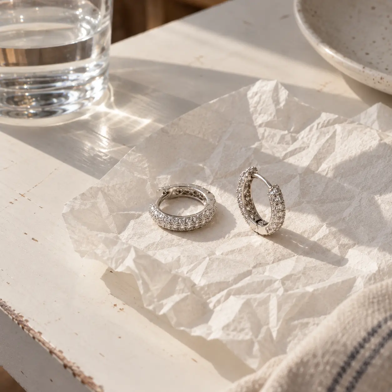 A pair of sparkling white-gold diamond huggie hoop earrings resting on crumpled white paper.